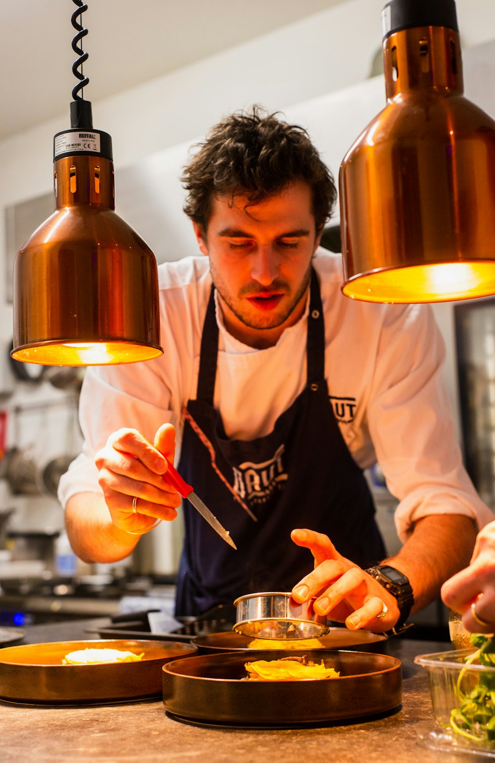A cook finishing plates under copper heat lamps at the pass
